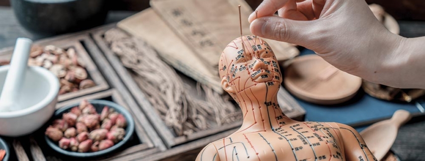 A hand inserts a needle into an acupuncture model surrounded by traditional Chinese medicine items