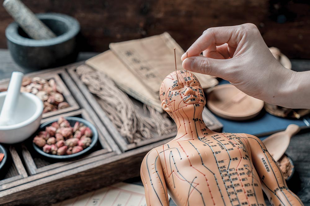 A hand inserts a needle into an acupuncture model surrounded by traditional Chinese medicine items